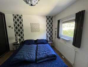 Ein Schlafzimmer mit einem Bett, einem Nachttisch, einer Lampe, einem Fenster mit Vorhängen und einer Heizung.