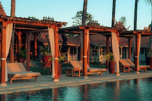 A poolside area with wooden lounge chairs and a covered seating area with a tiled roof.