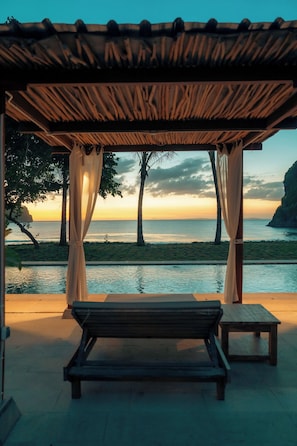 A beachside lounge with a wooden bench and a table, overlooking the sea and a sunset.