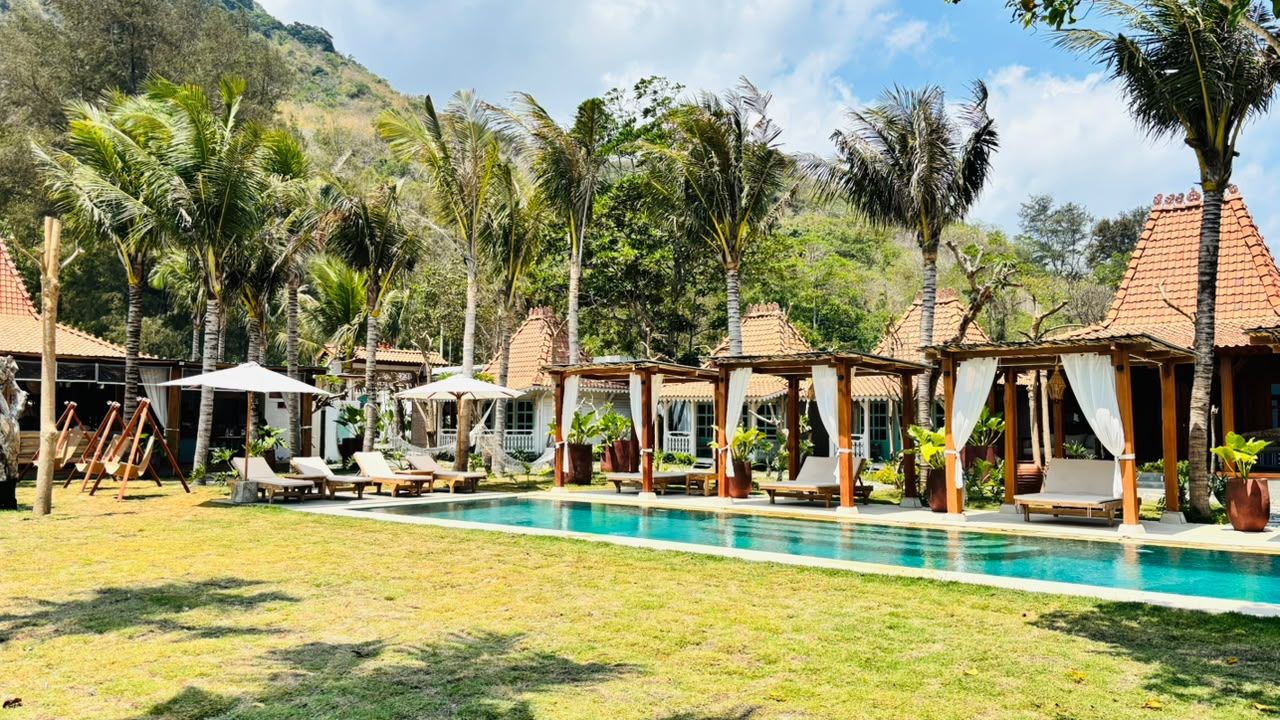 A pool area with lounge chairs and umbrellas, surrounded by palm trees and buildings with red-tiled roofs.