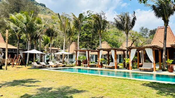 A pool area with lounge chairs and umbrellas, surrounded by palm trees and buildings with red-tiled roofs.