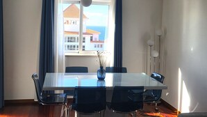 Ein Essbereich mit Tisch und Stühlen, einer Vase mit Trockenpflanzen, einem Fenster mit Vorhängen und Blick auf Gebäude und das Meer.