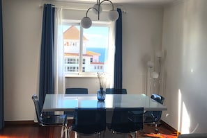 Uma sala de jantar com mesa e cadeiras, um vaso com plantas secas, uma janela com cortinas e vista para edifícios e o mar.