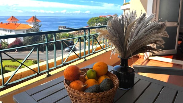 Ein Balkon mit einem Korbwäsche voller Früchte, einer Vase mit getrocknetem Pampasgras und Blick auf das Meer und Gebäude.