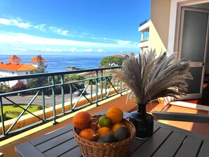 A balcony with a wicker basket of fruits, a vase with dried pampas grass, and a view of the ocean and buildings. - ATLANTIC PARADISE II (Caniço)