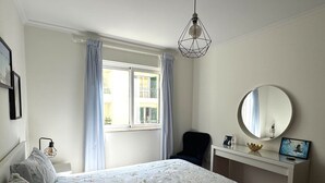A bedroom with a bed, a desk, a chair, a round mirror, and a window with curtains.