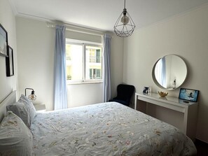 Quarto com cama, escrivaninha, cadeira, espelho redondo e janela com cortinas.