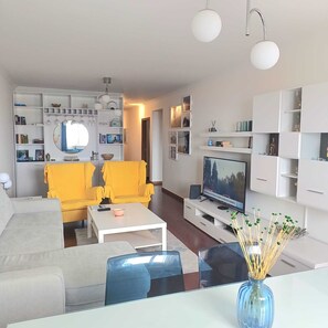 Uma sala de estar moderna com um sofá, cadeiras, uma mesa de centro, uma televisão e uma estante de livros.