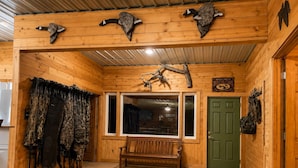 A cabin interior with wooden walls, a bench, hunting gear, and mounted animal heads.