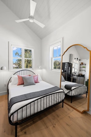 Ein Schlafzimmer mit Bett, Deckenventilator und Blick auf eine Küche durch ein Fenster.