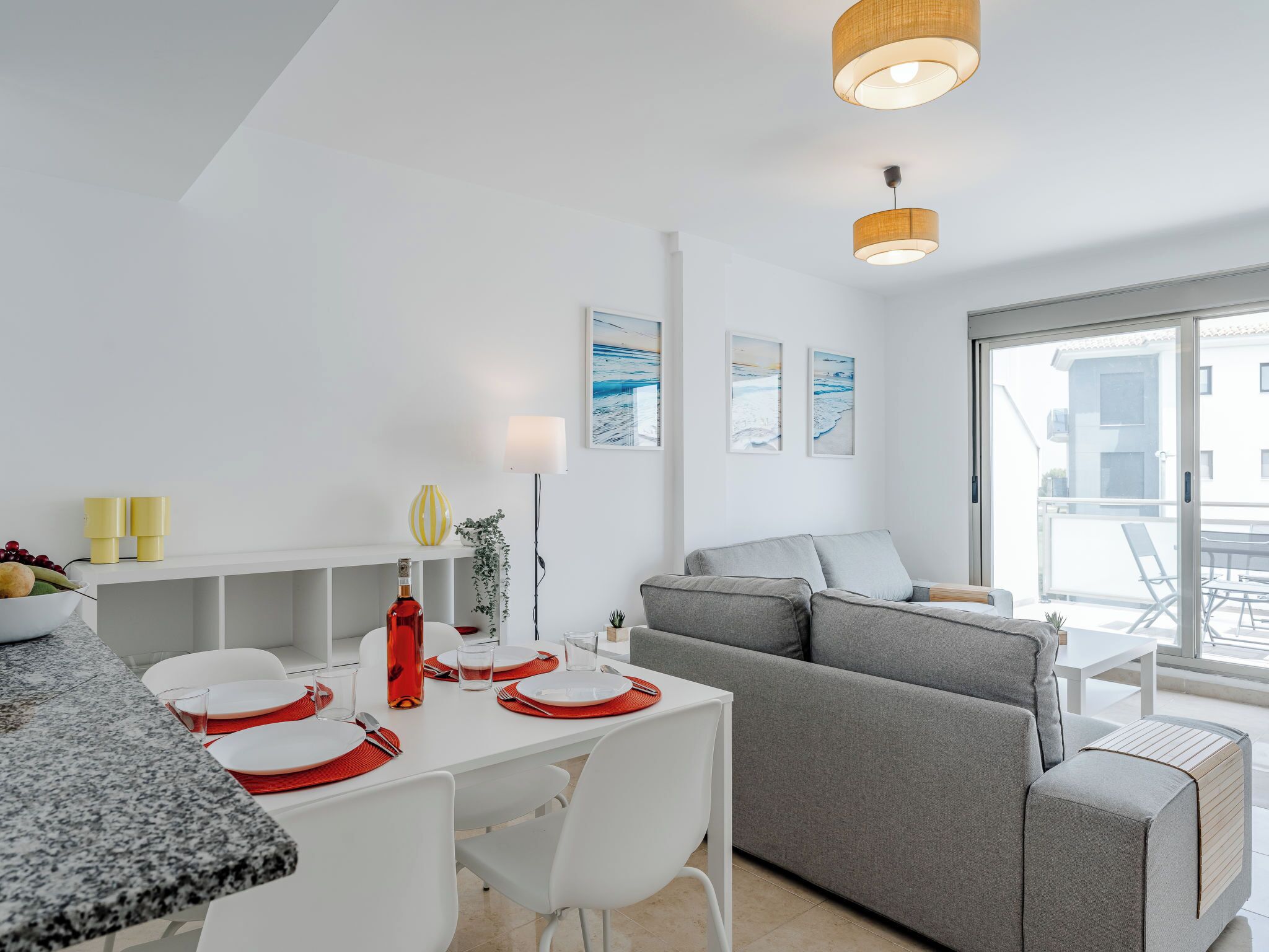 A modern, well-lit living area with a dining table set for two, a grey sofa, and a large window offering a view of the outside.