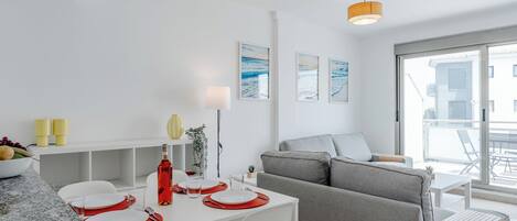 A modern, well-lit living area with a dining table set for two, a grey sofa, and a large window offering a view of the outside.