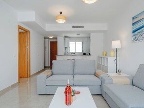A modern living room with a grey sectional sofa, a white coffee table with a bottle and glass, and a kitchen in the background. - Oliva Nova Cabagua by Interhome (Oliva)