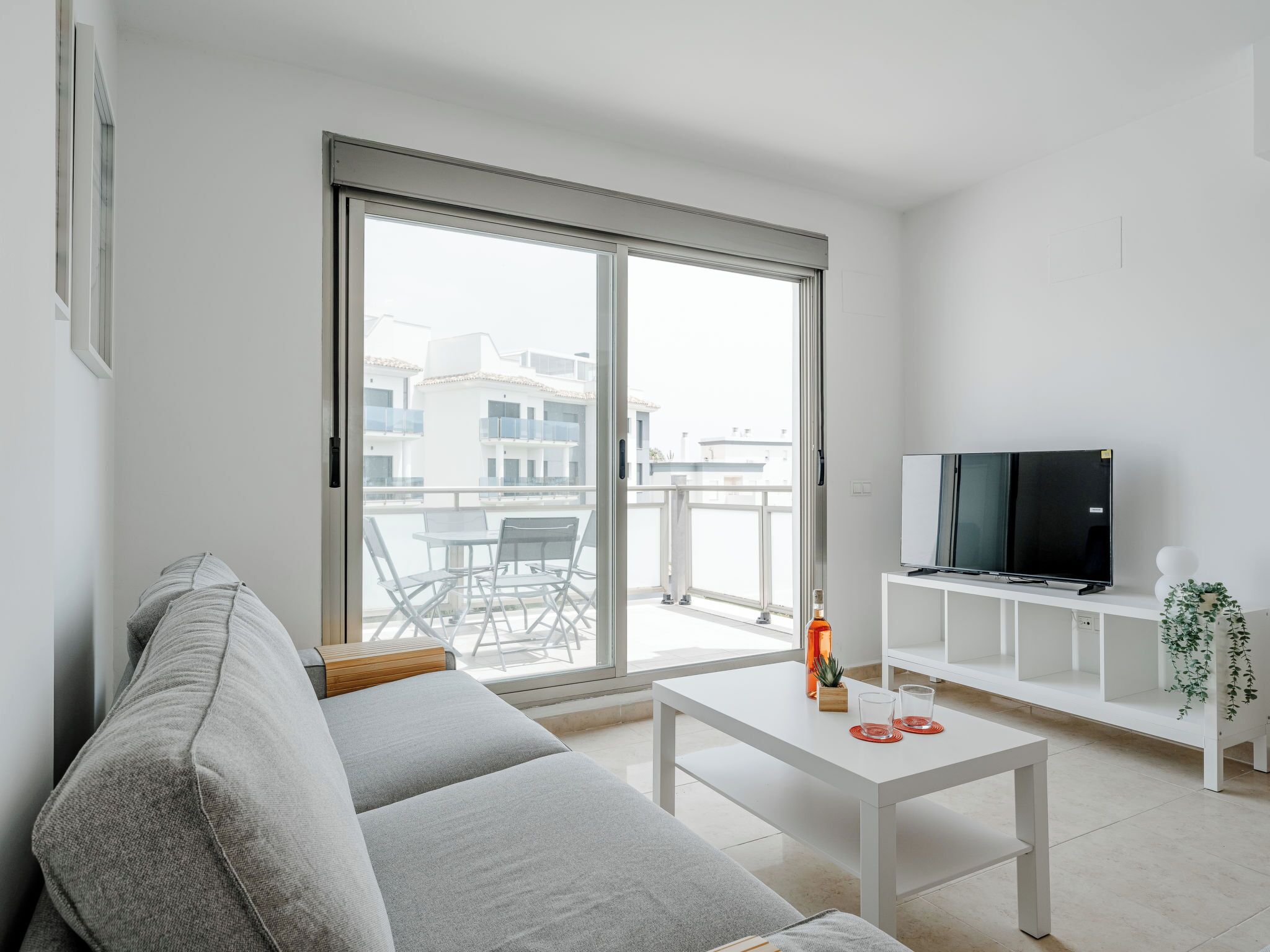 Un salón moderno con un sofá gris, una mesa de centro blanca, un televisor sobre un mueble blanco y un ventanal corredizo que da a un balcón exterior.