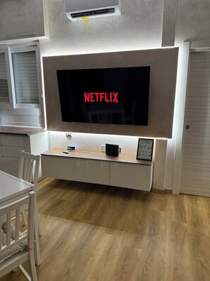 Ein modernes Wohnzimmer mit einem großen Flachbildfernseher an der Wand, einem Holzessttisch und einem weißen Sofa.