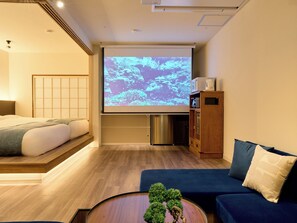 Hotellihuone, jossa on sänky, sohva, kahvinkeitin, projektorikangas ja pieni kaappi.