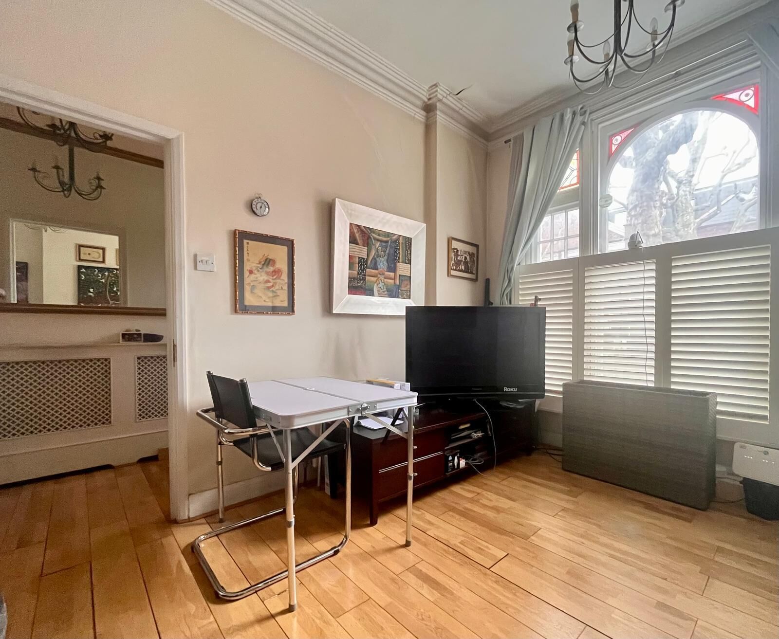 A living room with a flat-screen TV, a table, and a chair. There are framed pictures on the wall and a window with blinds.