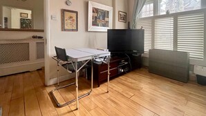 A living room with a flat-screen TV, a table, and a chair. There are framed pictures on the wall and a window with blinds. - Pay30nghts get up tp extra 59 free (Greater London)