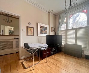 Ein Wohnzimmer mit Flachbildfernseher, Tisch und Stuhl. An der Wand befinden sich gerahmte Bilder und ein Fenster mit Jalousien.