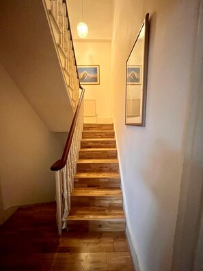 Eine Holztreppe mit weißen Geländern und einem Chandelier, das zu einer Zwischenebene führt, auf der ein Bild einer Berglandschaft gerahmt ist.