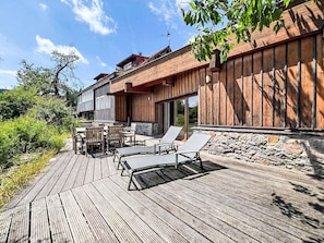 Eine Holzterrasse mit Gartenmöbeln, eine Steinmauer und ein Gebäude mit senkrechter Holzverkleidung.