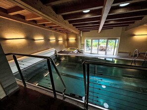 Ein Schwimmbecken mit Glasbarriere, Holzbalken an der Decke und Blick auf eine Terrasse.
