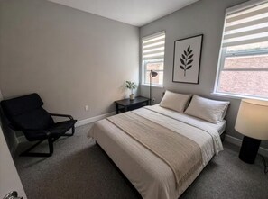 A bedroom with a bed, a chair, a nightstand, a lamp, and a window with blinds. - Breathtaking Modern Hillside Suite (San Jose)