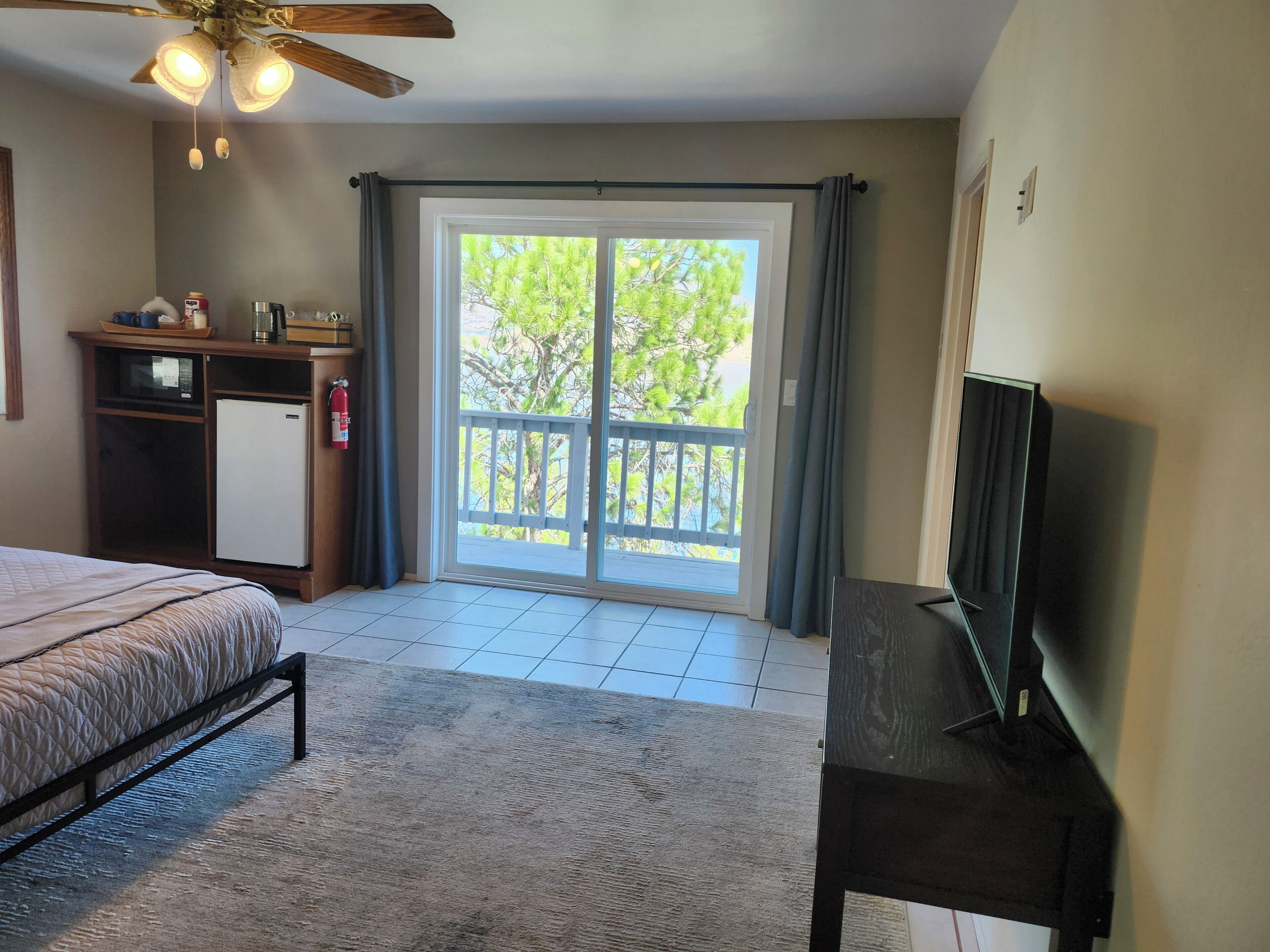 A bedroom with a bed, a desk, a television, a ceiling fan, and a balcony with a view of trees.