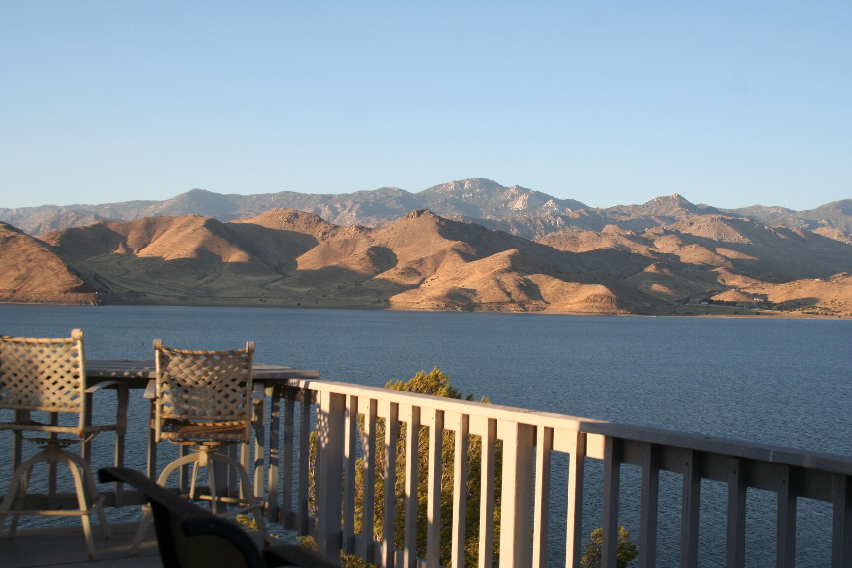 A lakeside terrace with chairs and a railing, overlooking a mountain range.