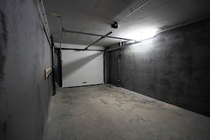Eine schlecht beleuchtete Garage mit einer weißen, geschlossenen Tür, Betonwänden und freiliegenden Rohren.