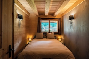 Ein gemütlicher Holzraum mit einem Bett, zwei Nachttischlampen, einem Fenster mit Blick auf Gebäude und Holzwänden.