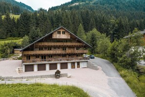 Eine Holzchalet mit Balkon, umgeben von einer Schotterauffahrt und einem bewaldeten Hang.