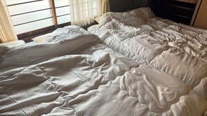 Uma cama com edredom e almofadas brancas, cabeceira de madeira e janela com cortinas.