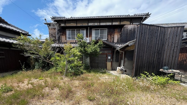 Uma casa japonesa tradicional com fachada de madeira, telhado com telhas e um pequeno jardim com grama e arbustos.