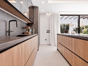 A modern kitchen with dark countertops, wooden cabinets, and a view of greenery through a window. - Villa Confort by Vacation Marbella (Estepona)