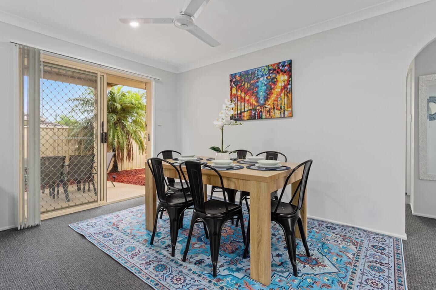 A dining area with a wooden table, black chairs, a colorful painting on the wall, and a patterned rug.