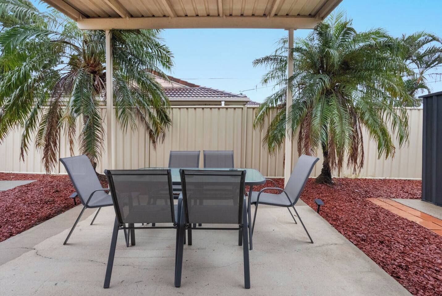 An outdoor patio with a table and chairs, surrounded by palm trees and a fence.