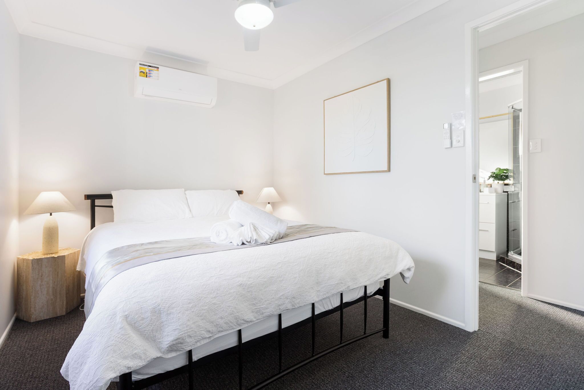 A neatly arranged bedroom with a bed, bedside lamps, a wall-mounted air conditioner, and a framed picture.