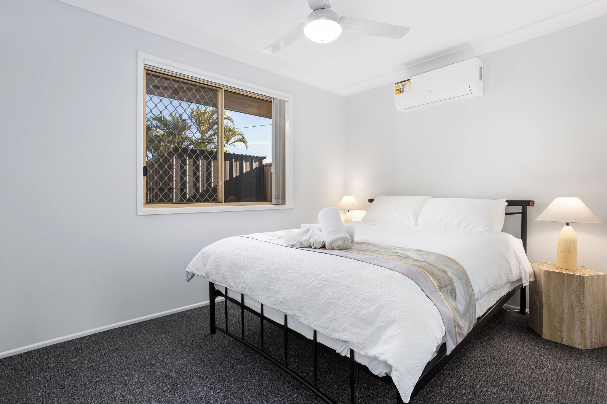A bedroom with a bed, two bedside lamps, a ceiling fan, and a window with blinds.