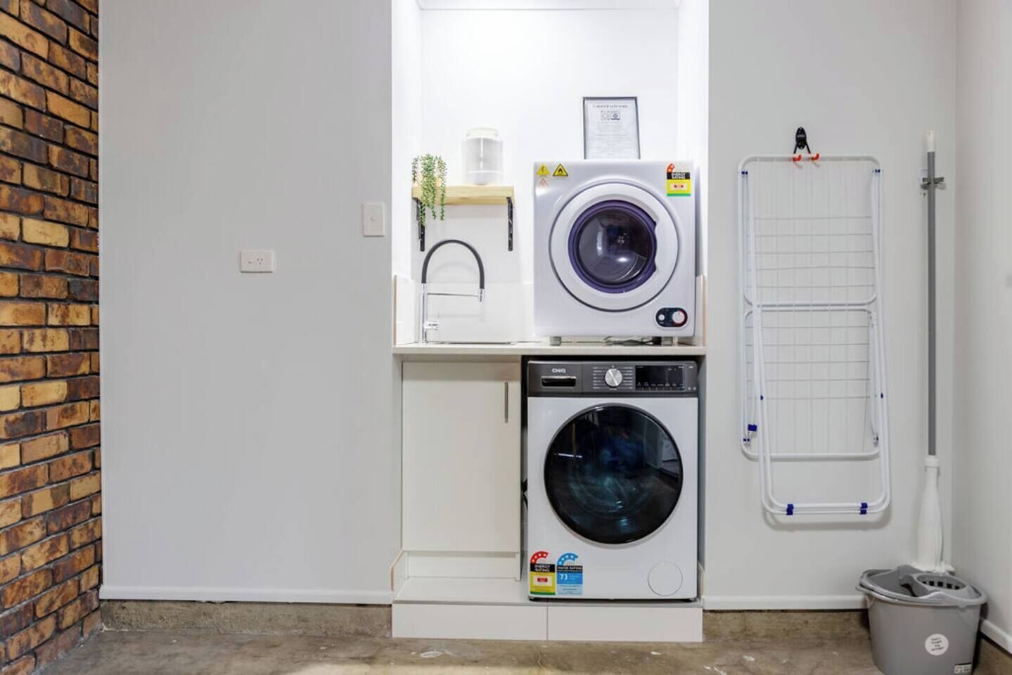 A laundry room with two washing machines, a drying rack, and a trash bin.