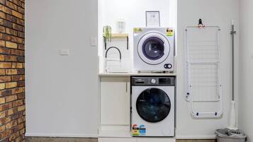 A laundry room with two washing machines, a drying rack, and a trash bin.