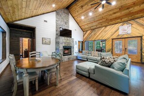 A spacious living area with a stone fireplace, wooden ceiling, and a large window overlooking greenery. - Sweet Magnolia Cabin with Hot Tub and Fire Pit (Broken Bow)