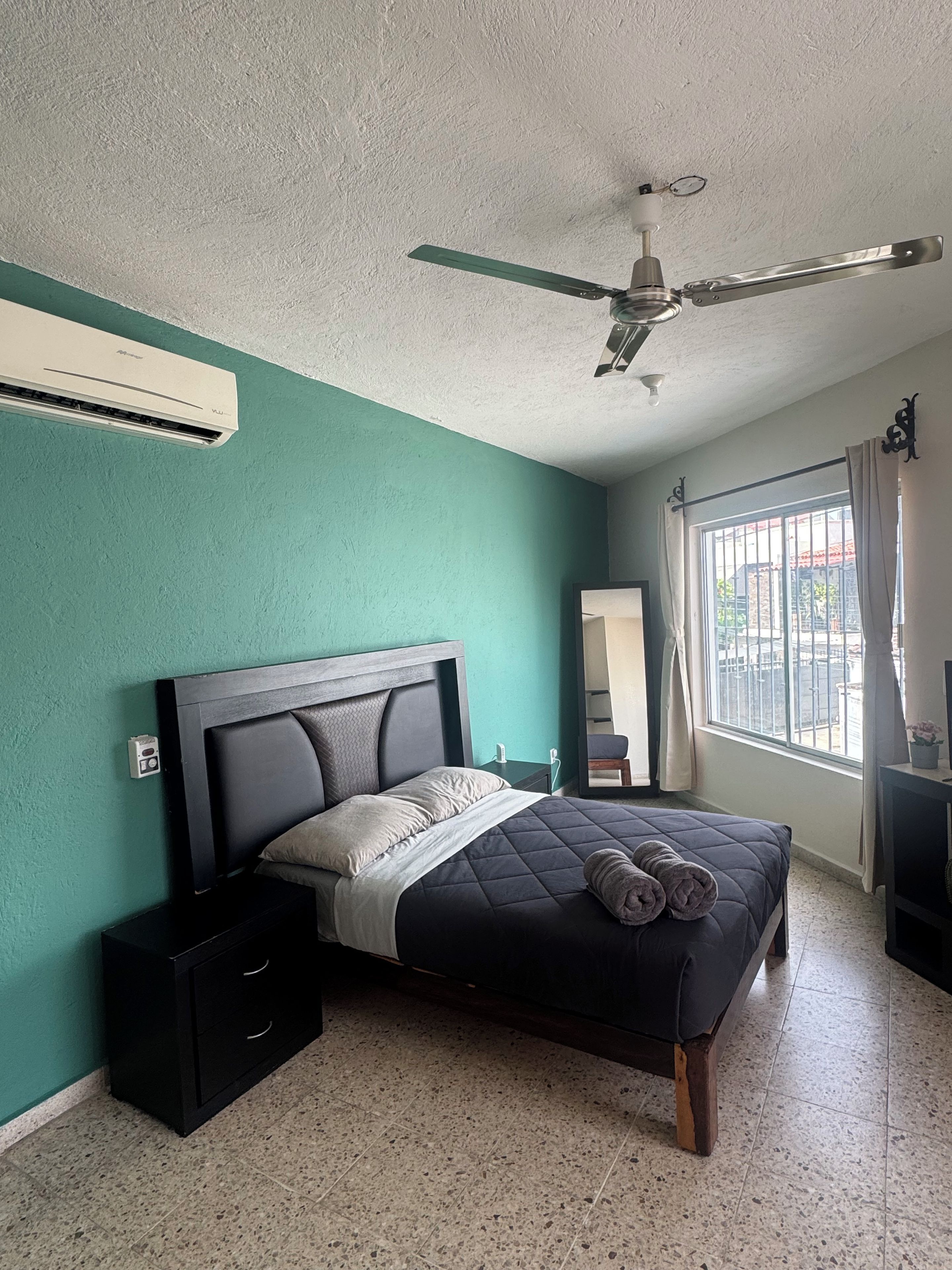 A bedroom with a bed, a ceiling fan, a window with curtains, and a wall-mounted air conditioner.