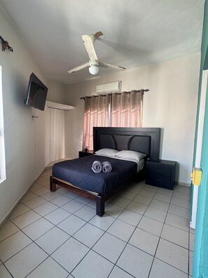Ein Schlafzimmer mit einem Bett, einem Nachttisch, einem Fernseher, einem Deckenventilator und einem Fenster mit Vorhängen.