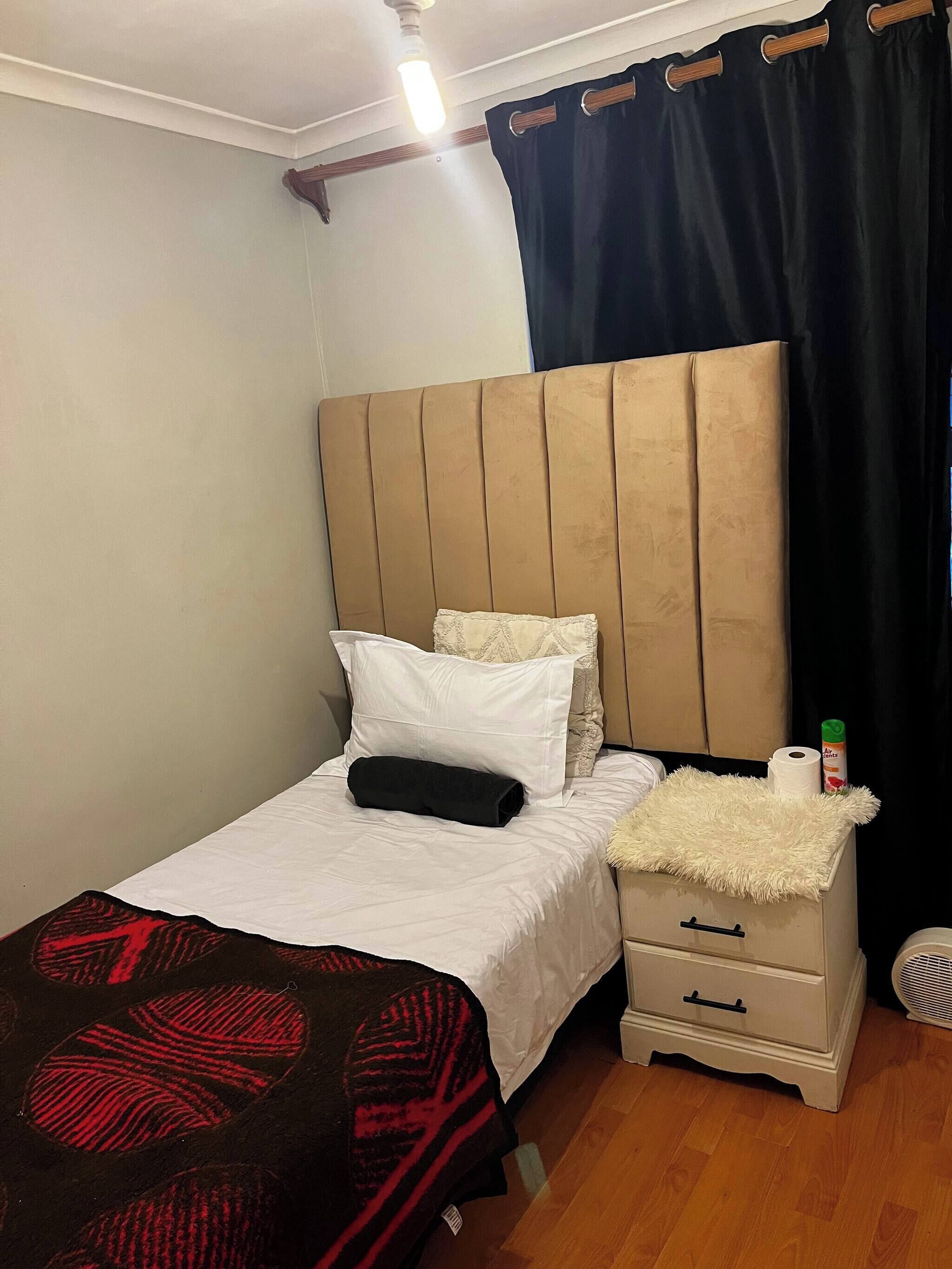 A single bed with a headboard, a bedside table with a drawer, a black and red blanket, and a small white nightstand with a bottle of air freshener.