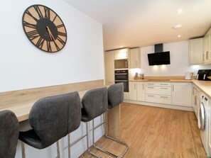 Een moderne keuken met een houten vloer, witte kastjes, een zwarte klok en een bar met stoelen.