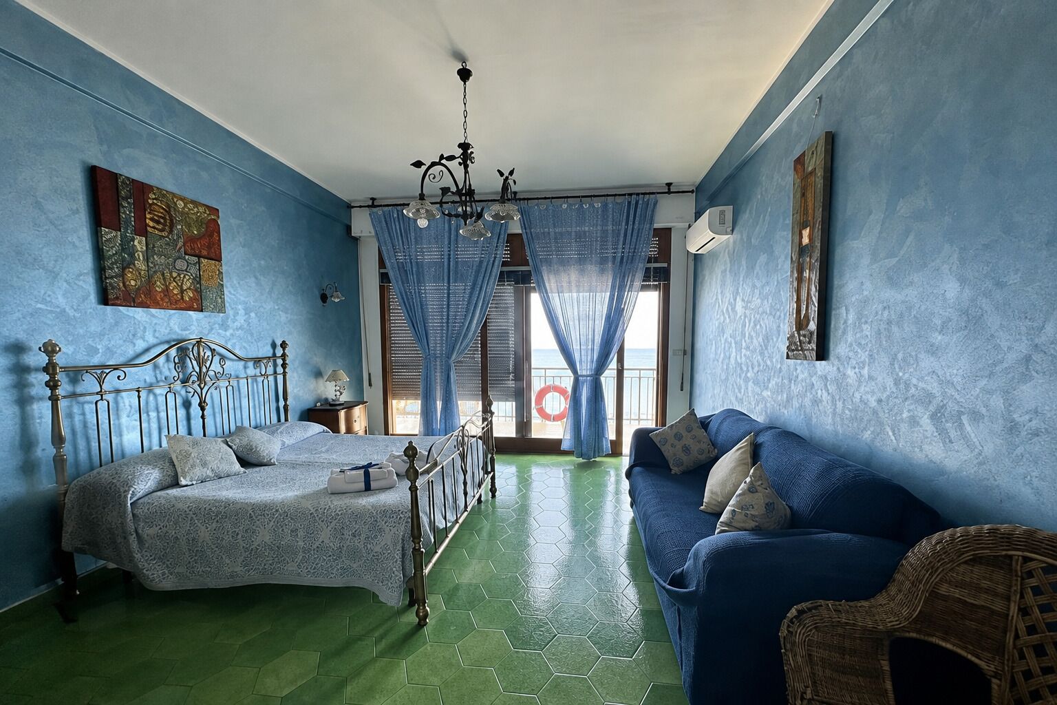 Una habitación con una cama, un sofá, una silla de mimbre, una araña y vista al mar a través de una ventana.