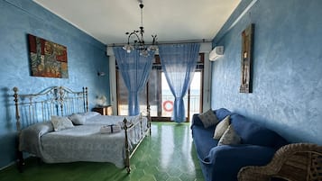 Una habitación con una cama, un sofá, una silla de mimbre, una araña y vista al mar a través de una ventana.