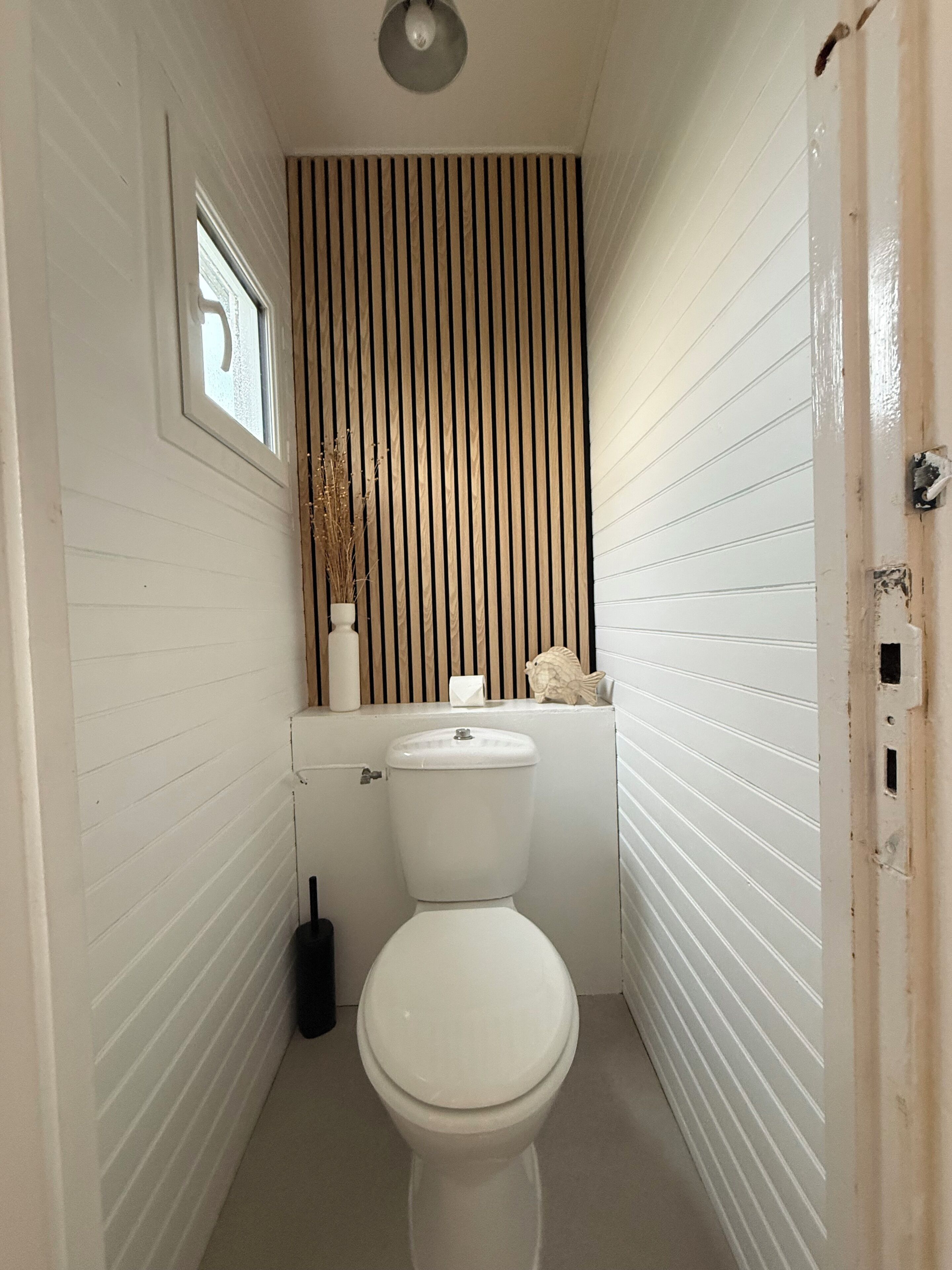 A small, narrow bathroom with a white toilet, a window, and a wooden slat wall.