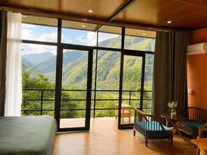 Habitación con un ventanal grande que ofrece vistas a la montaña, una cama, una silla de madera y una pequeña mesa con un florero.